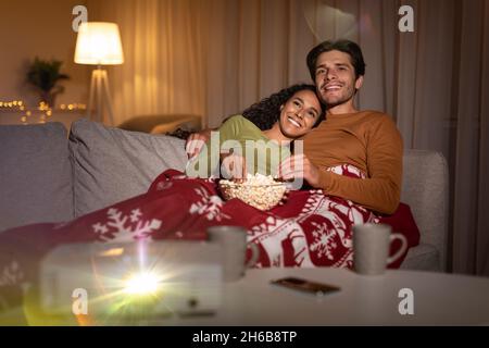 Film di Natale. Coppia felice famiglia guardando il film e mangiare Popcorn che giace avvolto in coperta sul divano a casa la vigilia di Natale. Sport invernali e divertimento Co Foto Stock