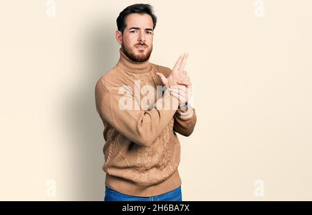 Giovane uomo ispanico che indossa abiti casual tenendo una pistola simbolica con gesto della mano, giocando uccidendo armi di tiro, faccia arrabbiata Foto Stock