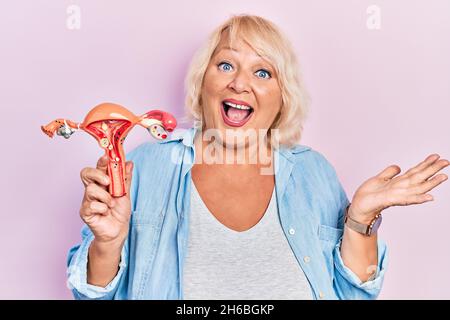 Donna bionda di mezza età che tiene il modello anatomico dell'organo genitale femminile che celebra la vittoria con sorriso felice e l'espressione del vincitore con le mani sollevate Foto Stock