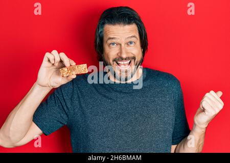 Medio età uomo caucasico mangiare proteine bar come snack energia sana puntando il pollice fino al lato sorridente felice con bocca aperta Foto Stock