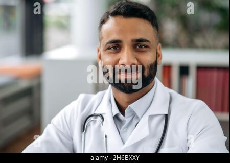 Primo piano ritratto di un generale indiano barbuto terapeuta, in uniforme medica e stetoscopio, seduto alla sua scrivania nel gabinetto, in ospedale, guardando la macchina fotografica, sorridente amichevole Foto Stock