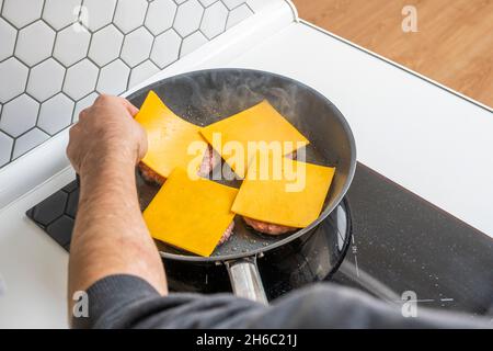 Mano dell'uomo che mette le fette di formaggio cheddar sugli hamburger di vitello alla griglia in una padella anti-scuotimento Foto Stock