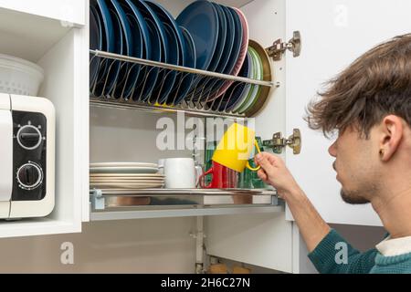 Giovane uomo che mette una tazza gialla nel mobile della cucina per scolare i piatti Foto Stock