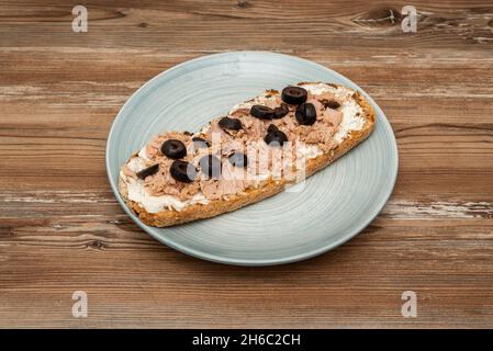 Brindisi di tonno in scatola con formaggio fresco spalmato con fette di olive nere su un piatto blu Foto Stock