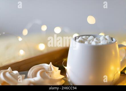 Cacao in una tazza su un vassoio di marshmallows e meringa. Luci di Capodanno sullo sfondo. Foto Stock