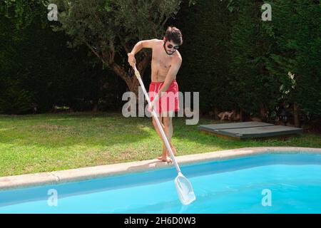 Giovane uomo che pulisce la piscina di foglie autunnali con rete di pulizia. Foto Stock