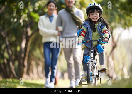 bambina asiatica con casco e completo di protezione in bicicletta nel parco della città con i genitori che guardano da dietro Foto Stock