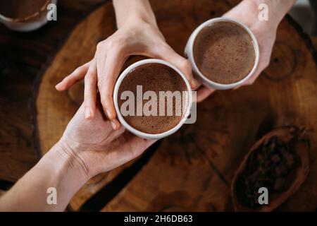 Cacao cerimoniale caldo fatto a mano in tazze bianche. Donna mani che danno cacao artigianale, vista dall'alto su tavola di legno. Bevanda biologica sana al cioccolato preparata da Foto Stock