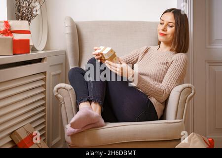 BELLA giovane donna mentre si siede in una comoda poltrona che tiene una scatola regalo circondata da regali a casa. GHE intuisce cosa c'è dentro Foto Stock