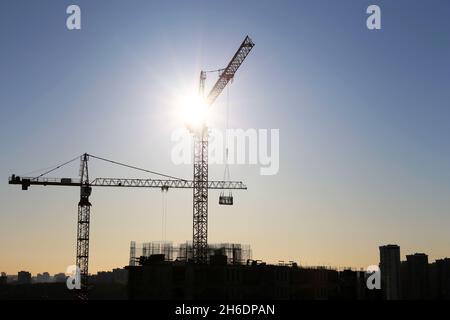 Sagome di gru edili ed edifici residenziali incompleti su sfondo solare. Edilizia abitativa, condominio in città Foto Stock