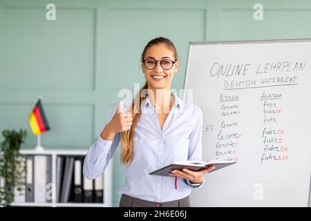 Allegra tutor femminile che consiglia la scuola di lingue straniere, mostrando pollice in su, dando la lezione di tedesco e sorridendo Foto Stock