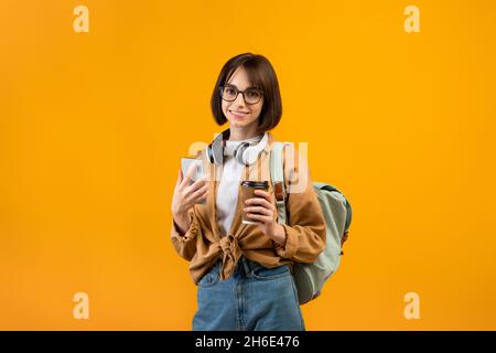 Positiva studentessa con smartphone, zaino e cuffie sul collo, caffè, sfondo giallo Foto Stock