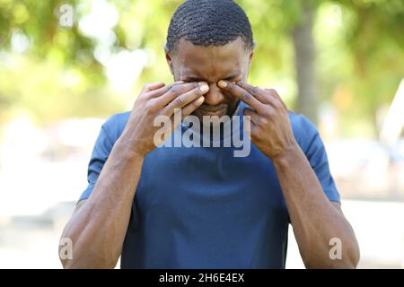 Ritratto della vista frontale di un uomo con la pelle nera che graffia gli occhi itchy in un parco Foto Stock