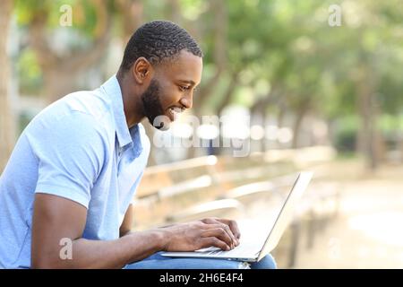 Ritratto laterale di un uomo felice con pelle nera utilizzando un computer portatile seduto su una panca in un parco Foto Stock