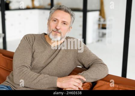 Bell'uomo maturo dai capelli grigi con una barba guarda la fotocamera. Testa di carismatico uomo anziano in casual vestirsi seduto in posa rilassata sul divano a casa Foto Stock