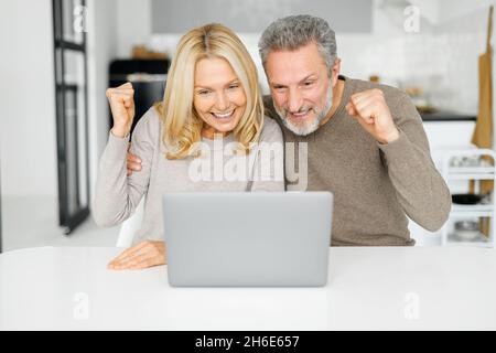 Overjoyed coppia di mezza età vincere nella lotteria in linea, ha ricevuto buone notizie, marito maturo felice e moglie che staring allo schermo del portatile e lo schermo sì felicemente, alzando la mano nel trionfo e nella vittoria Foto Stock