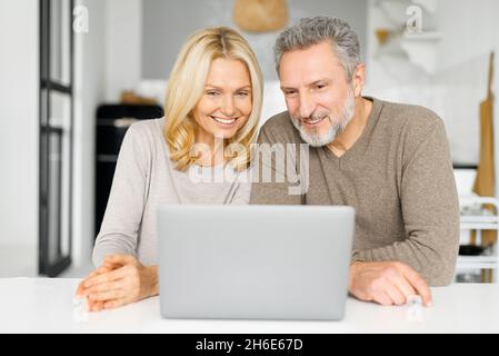 Una coppia felice di mezza età trascorre del tempo libero con un computer portatile a casa. Gli sposi senior si siedono alla scrivania in cucina, guardando video, navigare sul Web o fare shopping online insieme, guardare lo schermo e sorridere Foto Stock