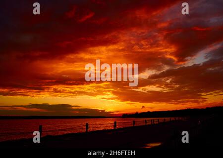 Cielo sanguinoso sopra il ponte e onde superficiali riflettenti dell'acqua. Rosso tramonto nuvoloso sopra il lago al crepuscolo. Nuvolosità serale al crepuscolo. Cielo ruddy orizzonte Foto Stock