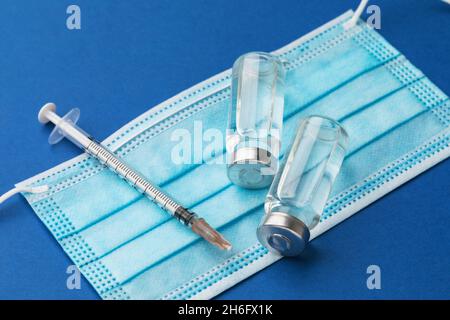Siringa monouso, soluzione per iniezione e maschera medica su sfondo blu, concetto sul tema della vaccinazione contro il coronavirus Foto Stock