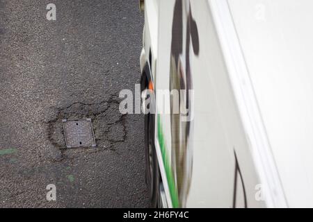 I veicoli passano un buco della pentola e la superficie della strada danneggiata a Godley Cutting, Halifax, West Yorkshire, Regno Unito. Una pothole è una depressione in una superficie stradale, di solito asfalto pavimentazione, dove il traffico ha rimosso pezzi rotti del marciapiede. È solitamente il risultato di acqua nella struttura di suolo sottostante e traffico che passa sopra l'area colpita. L'acqua prima indebolisce il terreno sottostante; il traffico poi si affatica e rompe la superficie di asfalto mal sostenuta nell'area colpita. L'azione continua del traffico espelle sia l'asfalto che il materiale del terreno sottostante per creare un buco nel marciapiede. Foto Stock