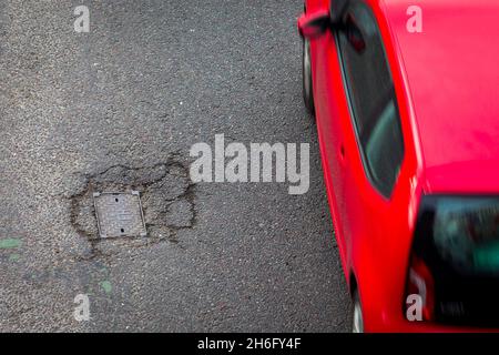 I veicoli passano un buco della pentola e la superficie della strada danneggiata a Godley Cutting, Halifax, West Yorkshire, Regno Unito. Una pothole è una depressione in una superficie stradale, di solito asfalto pavimentazione, dove il traffico ha rimosso pezzi rotti del marciapiede. È solitamente il risultato di acqua nella struttura di suolo sottostante e traffico che passa sopra l'area colpita. L'acqua prima indebolisce il terreno sottostante; il traffico poi si affatica e rompe la superficie di asfalto mal sostenuta nell'area colpita. L'azione continua del traffico espelle sia l'asfalto che il materiale del terreno sottostante per creare un buco nel marciapiede. Foto Stock