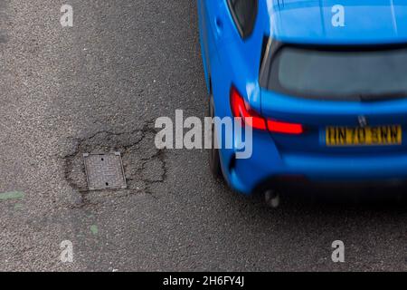 I veicoli passano un buco della pentola e la superficie della strada danneggiata a Godley Cutting, Halifax, West Yorkshire, Regno Unito. Una pothole è una depressione in una superficie stradale, di solito asfalto pavimentazione, dove il traffico ha rimosso pezzi rotti del marciapiede. È solitamente il risultato di acqua nella struttura di suolo sottostante e traffico che passa sopra l'area colpita. L'acqua prima indebolisce il terreno sottostante; il traffico poi si affatica e rompe la superficie di asfalto mal sostenuta nell'area colpita. L'azione continua del traffico espelle sia l'asfalto che il materiale del terreno sottostante per creare un buco nel marciapiede. Foto Stock