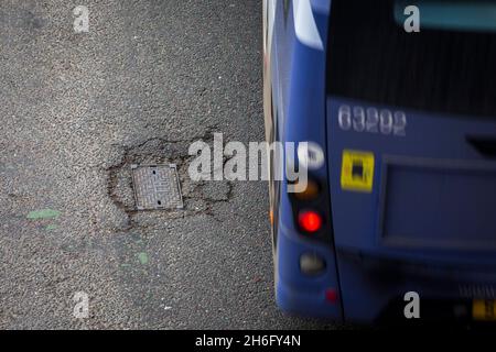 I veicoli passano un buco della pentola e la superficie della strada danneggiata a Godley Cutting, Halifax, West Yorkshire, Regno Unito. Una pothole è una depressione in una superficie stradale, di solito asfalto pavimentazione, dove il traffico ha rimosso pezzi rotti del marciapiede. È solitamente il risultato di acqua nella struttura di suolo sottostante e traffico che passa sopra l'area colpita. L'acqua prima indebolisce il terreno sottostante; il traffico poi si affatica e rompe la superficie di asfalto mal sostenuta nell'area colpita. L'azione continua del traffico espelle sia l'asfalto che il materiale del terreno sottostante per creare un buco nel marciapiede. Foto Stock