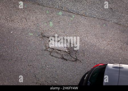 I veicoli passano un buco della pentola e la superficie della strada danneggiata a Godley Cutting, Halifax, West Yorkshire, Regno Unito. Una pothole è una depressione in una superficie stradale, di solito asfalto pavimentazione, dove il traffico ha rimosso pezzi rotti del marciapiede. È solitamente il risultato di acqua nella struttura di suolo sottostante e traffico che passa sopra l'area colpita. L'acqua prima indebolisce il terreno sottostante; il traffico poi si affatica e rompe la superficie di asfalto mal sostenuta nell'area colpita. L'azione continua del traffico espelle sia l'asfalto che il materiale del terreno sottostante per creare un buco nel marciapiede. Foto Stock