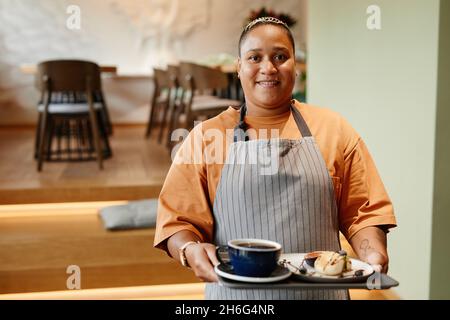 Ritratto orizzontale medio di giovane donna che lavora in caffè portando caffè e dessert ordinato al cliente Foto Stock