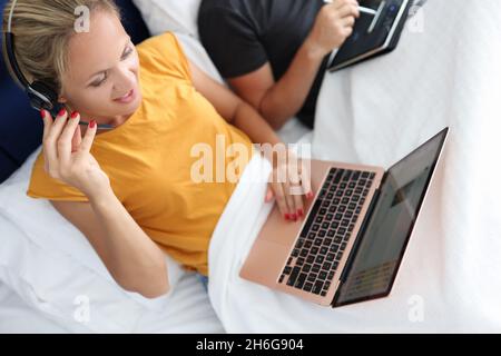 La giovane donna in cuffie con microfono funziona su un computer portatile mentre si trova a letto Foto Stock