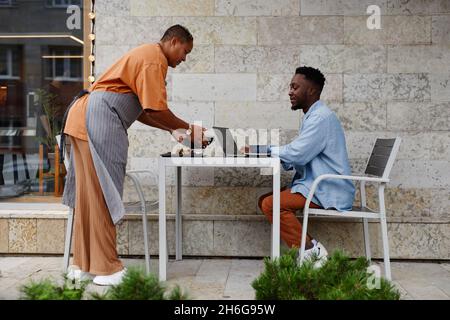 Vista laterale di un giovane uomo afroamericano che indossa abiti casual seduti al tavolo da caffè all'aperto, cameriera che porta cibo ordinato a lui Foto Stock
