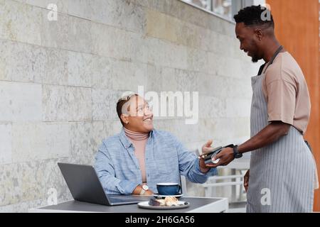 Gioiosa giovane donna nera seduta al tavolo all'aperto caffè a pagamento per il pranzo con il suo smartphone Foto Stock