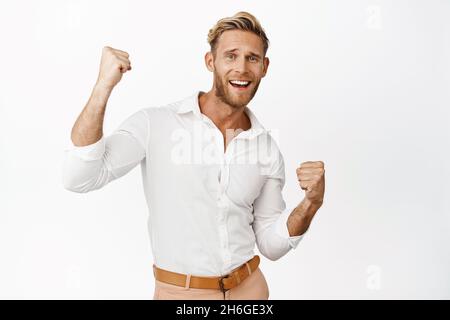 Ritratto dell'uomo fortunato vincitore che festeggia, alza i pugni, si sente entusiasta e festeggia, in piedi su sfondo bianco Foto Stock