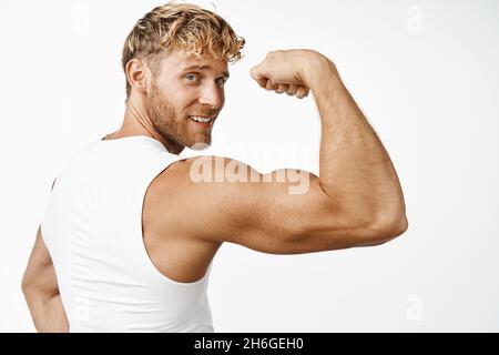 Sportivo flessibile bicipite, mostrando braccio forte e sorridente felice dopo l'allenamento in palestra, sfondo bianco Foto Stock