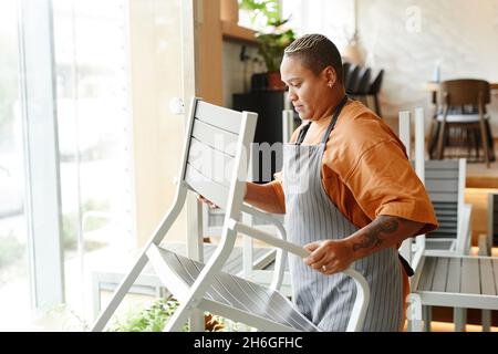 Vista laterale di giovane donna elegante che lavora in moderno caffè mettendo sedie e tavoli prima di aprire Foto Stock