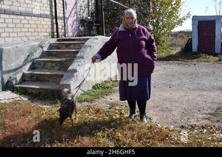 Verkhnotoretske, Donetsk, Ucraina. 16 ottobre 2021. Una signora anziana si alza con il suo cane sul retro della sua casa nella periferia del villaggio di Verkhnotoretske. Verkhnotoretske è un insediamento ucraino di tipo urbano situato in prima linea nel distretto di Yasynuvata, nell'oblast di Donetsk dell'Ucraina orientale, a 24 km dal centro della città di Donetsk (DPR). Durante la guerra di Donbas, iniziata a metà aprile 2014, la linea di separazione tra le parti in guerra è stata situata nelle vicinanze dell'insediamento. Il villaggio era una zona grigia fino alla fine del 2017 anno. Al momento Verkhnotoretske Foto Stock