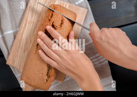 Vista dall'alto delle mani della donna che tengono il coltello e tagliano il pane fresco della pasta madre sul tagliere Foto Stock