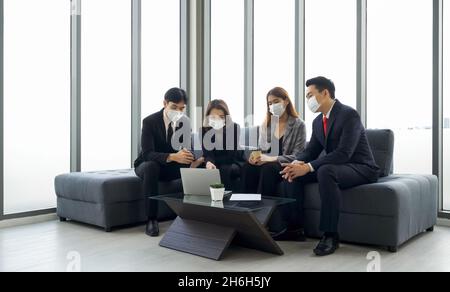 Gruppo di uomini d'affari in tuta discutere dei dati sul computer portatile. Tutti indossano una maschera. Concetto di team aziendale di persone. Foto Stock