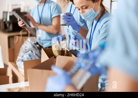 Primo piano di volontari impegnati indossando uniformi blu, maschere protettive e guanti di smistamento donato cibo oggetti mentre si lavora in fondazione beneficenza Foto Stock