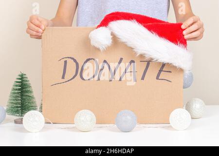 Mani che tengono un pezzo di cartone marrone con iscrizione Donate e Babbo Natale cappello. Donazione di Natale, concetto di carità vacanza Foto Stock