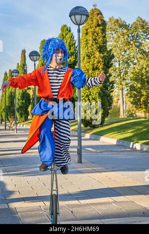 Pieno corpo di uomo divertente in costume clown e parrucca, giraffa uniciclo con braccia distese su sentiero lastricato in parco verde in giorno di sole Foto Stock