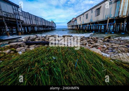 Due vecchi moli di legno sporgono nel mare di fronte a rocce, flora e spazzatura lavata in Hua Hin, Thailandia Foto Stock