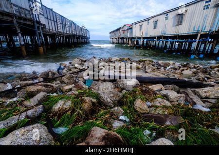 Due vecchi moli di legno sporgono nel mare di fronte a rocce, flora e spazzatura lavata in Hua Hin, Thailandia Foto Stock