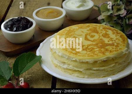 Frittelle russe crepes in pila e sulla padella con salsa di mele e panna acida su sfondo rustico in legno. Pila di pancake blini russi su un legno Foto Stock