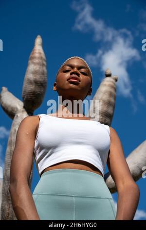 Fidata atleta afro-americana in abbigliamento sportivo e con capelli corti in piedi contro l'albero drago nel parco e guardando la macchina fotografica Foto Stock