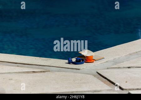 coppa d'arancia, libro, auricolari blu vicino alla piscina. Foto di alta qualità Foto Stock