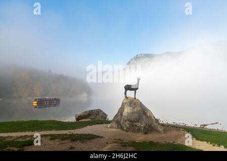 Bohinj, Slovenia - 19 ottobre 2021: Statua di un camoscio su una roccia sul lago di Bohinj in una mattinata d'autunno nebbia con una barca di legno sullo sfondo Foto Stock