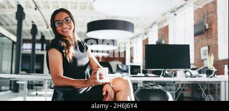donna d'affari che fa una pausa caffè al lavoro. Giovane donna d'affari felice sorridente allegramente mentre tiene in mano una tazza di caffè. Giovane donna d'affari seduta a Foto Stock