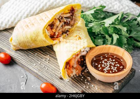 Rotolo di shawarma con semi di sesamo, salsa barbecue, pollo e verdure in pita pane su sfondo legno scuro con pomodori ciliegini e pars verdi Foto Stock