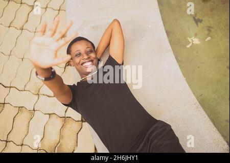 Primo piano ritratto di attraente felice sorridente giovane bellezza naturale capelli corti donna africana che indossa nero t-shirt ridere allungando le mani di chiusura dalla macchina fotografica che si trova sul piano terra vista Foto Stock